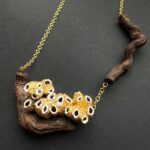 Collar creado a partir de la raiz de una cepa de los viñedos del Penedés seccionada en dos partes que se unen por la misma cadena que sostiene el collar en acero inoxidable bañado en oro de 18 k. Sobre la raiz reposa una familia de barnacles naturales recolectados en las playas del Garraf y pintados en dorado y bocas blancas.